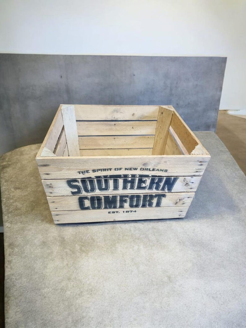 Pode incluir: Uma caixa de madeira com as palavras "SOUTHERN COMFORT" em letras pretas. O texto "THE SPIRIT OF NEW ORLEANS" e "EST. 1874" tamb&eacute;m s&atilde;o vis&iacute;veis. A caixa &eacute; feita de madeira clara e est&aacute; sobre uma superf&iacute;cie cinza.