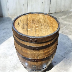 May include: A wooden barrel with a flat top and metal bands. The barrel is made of light brown wood with a natural grain pattern. The metal bands are dark brown and encircle the barrel. The top of the barrel is also made of wood.