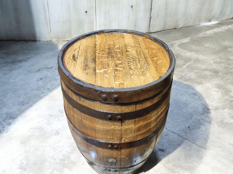May include: A wooden barrel with a flat top and metal bands. The barrel is made of light brown wood with a natural grain pattern. The metal bands are dark brown and encircle the barrel. The top of the barrel is also made of wood.