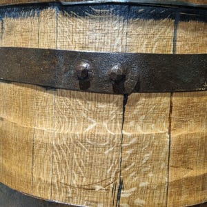 May include: Close-up of a wooden barrel with dark metal bands and rivets. The wood grain is visible, with dark staining between the staves. The metal bands are riveted to the wood, creating a rustic appearance.