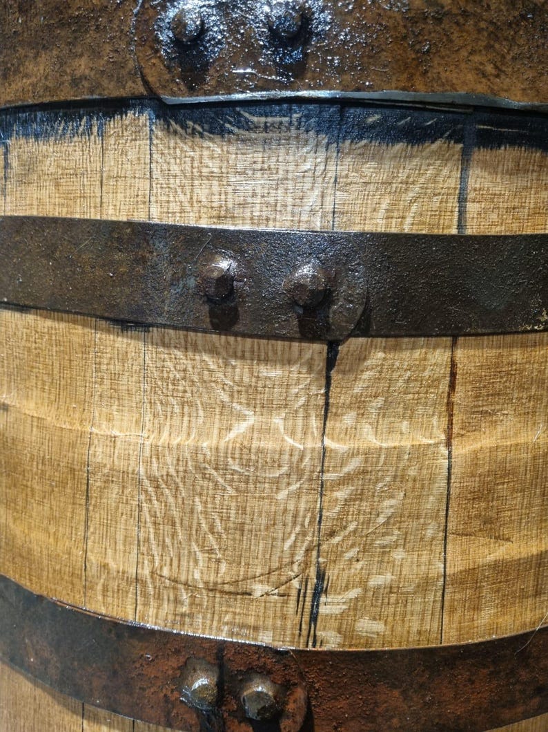 May include: Close-up of a wooden barrel with dark metal bands and rivets. The wood grain is visible, with dark staining between the staves. The metal bands are riveted to the wood, creating a rustic appearance.