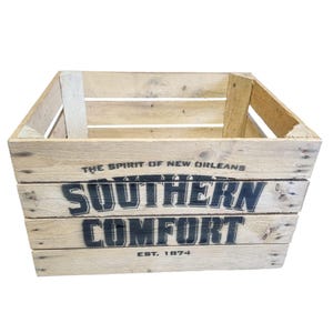 Pode incluir: Caixa de madeira com as palavras "SOUTHERN COMFORT" em letras pretas. A caixa &eacute; feita de madeira clara e tem o texto "THE SPIRIT OF NEW ORLEANS" e "EST. 1874" impresso.