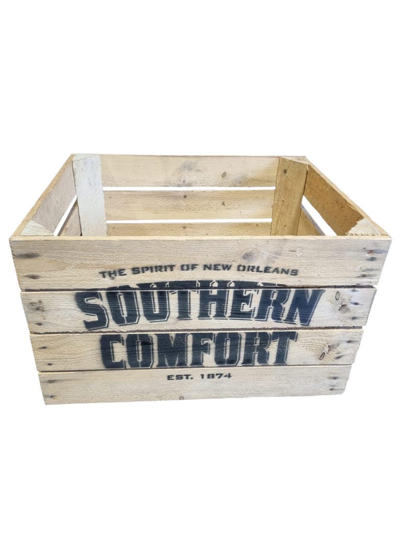 Pode incluir: Caixa de madeira com as palavras "SOUTHERN COMFORT" em letras pretas. A caixa &eacute; feita de madeira clara e tem o texto "THE SPIRIT OF NEW ORLEANS" e "EST. 1874" impresso.