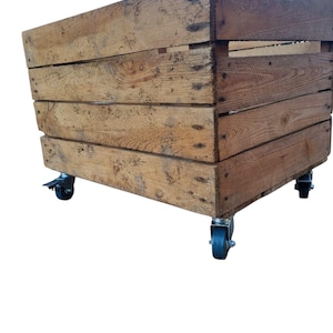 May include: A rustic, rectangular wooden crate with visible wood grain and nail heads. The crate is mounted on four black caster wheels, allowing for easy movement. The wood has a natural, weathered appearance.
