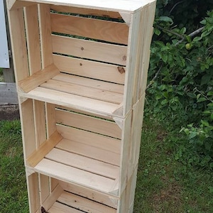 May include: A three-tier wooden shelf unit made from natural wood crates. The shelf unit is made of three stacked crates, each with a slatted front and sides. The shelf unit is standing on a grassy surface.
