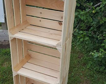 May include: A three-tier wooden shelf unit made from natural wood crates. The shelf unit is made of three stacked crates, each with a slatted front and sides. The shelf unit is standing on a grassy surface.