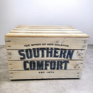 Pode incluir: Uma caixa de madeira com o texto "SOUTHERN COMFORT" em letras pretas. A caixa &eacute; feita de madeira clara e tem a frase "THE SPIRIT OF NEW ORLEANS" e "EST. 1874" impressas.