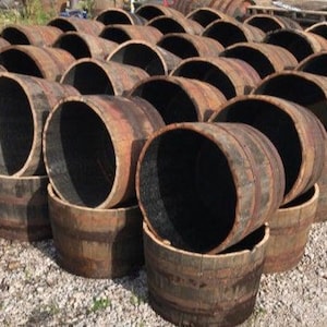 Può includere: Una collezione di botti e mezze botti di legno stagionato, disposte in file. Le botti sono marroni con interni scuri e cerchi metallici. La scena è all'aperto su una superficie di ghiaia.