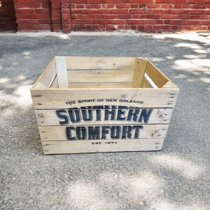 Pode incluir: Caixa de madeira clara com o texto "SOUTHERN COMFORT" em preto, com "EST. 1874" abaixo. A caixa &eacute; feita de madeira clara e tem formato retangular. A frase "THE SPIRIT OF NEW ORLEANS" est&aacute; acima do texto principal.