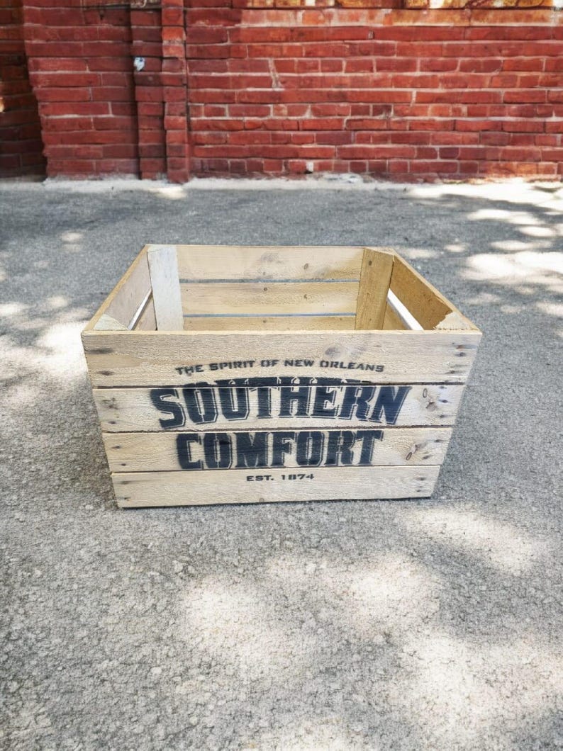 Pode incluir: Caixa de madeira clara com o texto "SOUTHERN COMFORT" em preto, com "EST. 1874" abaixo. A caixa &eacute; feita de madeira clara e tem formato retangular. A frase "THE SPIRIT OF NEW ORLEANS" est&aacute; acima do texto principal.