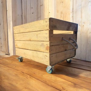 May include: A rustic wooden storage crate with a natural finish. The crate is rectangular, constructed from horizontal wooden planks, and features rope handles. It is mounted on four small caster wheels for easy mobility.
