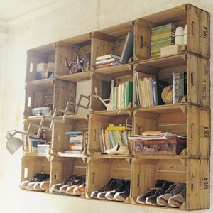 May include: A wall unit made of wooden crates, stacked and arranged to create shelves. The crates are painted in a natural wood color and have the year "1954" printed on them. The shelves are filled with books, shoes, and other items.