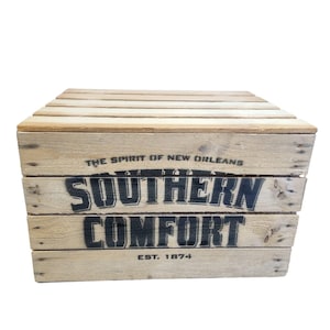Pode incluir: Uma caixa de madeira com o texto "SOUTHERN COMFORT" em preto, juntamente com "THE SPIRIT OF NEW ORLEANS" e "EST. 1874". A caixa &eacute; feita de madeira clara.
