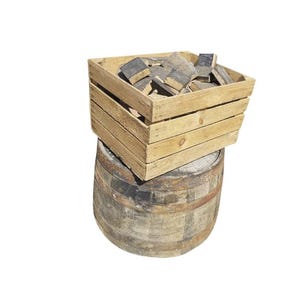 Op de afbeelding: Een lichtgekleurde houten krat gevuld met kleine, rechthoekige, verkoold houten blokjes, rustend op een grote, verweerde, cilindrische houten ton met donkere metalen banden en een rustieke, getextureerde afwerking.