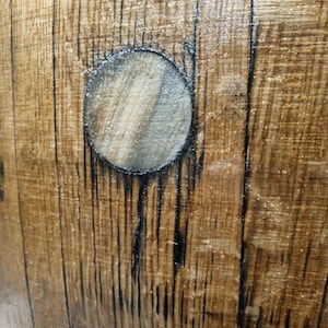 May include: Close-up of a section of a wooden barrel. The wood has a warm brown tone with dark vertical lines and a circular opening. The wood grain is visible, and the surface has a glossy finish.