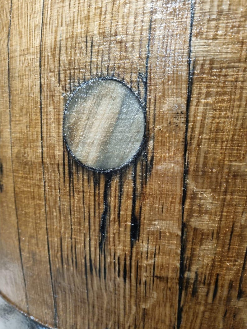 May include: Close-up of a section of a wooden barrel. The wood has a warm brown tone with dark vertical lines and a circular opening. The wood grain is visible, and the surface has a glossy finish.