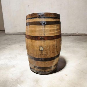 May include: A wooden barrel with metal bands. The barrel is made of light brown wood and has several dark brown metal bands wrapped around it. The barrel is standing upright on a concrete floor.