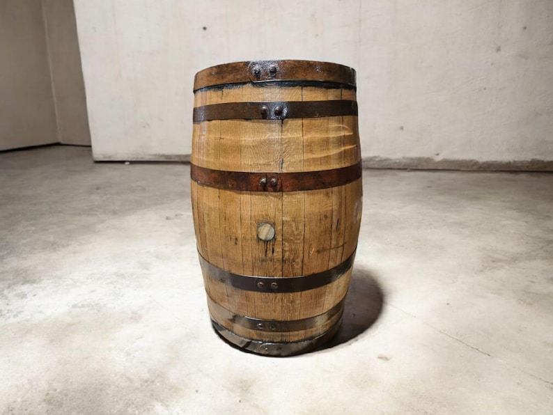 May include: A wooden barrel with metal bands. The barrel is made of light brown wood and has several dark brown metal bands wrapped around it. The barrel is standing upright on a concrete floor.