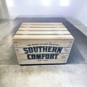 Pode incluir: Uma caixa de madeira com as palavras "SOUTHERN COMFORT" em letras pretas. A caixa &eacute; feita de madeira clara e tem o texto "THE SPIRIT OF NEW ORLEANS" e "EST. 1874" impresso. A caixa est&aacute; em um ch&atilde;o de concreto.