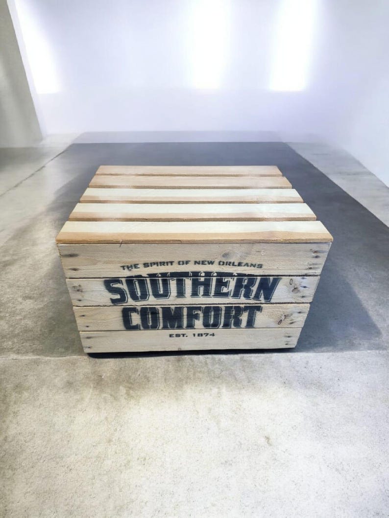 Pode incluir: Uma caixa de madeira com as palavras "SOUTHERN COMFORT" em letras pretas. A caixa &eacute; feita de madeira clara e tem o texto "THE SPIRIT OF NEW ORLEANS" e "EST. 1874" impresso. A caixa est&aacute; em um ch&atilde;o de concreto.