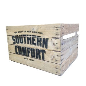 Pode incluir: Uma caixa de madeira r&uacute;stica com as palavras "SOUTHERN COMFORT" em letras pretas grandes. O texto "THE SPIRIT OF NEW ORLEANS" e "EST. 1874" tamb&eacute;m s&atilde;o vis&iacute;veis. A caixa &eacute; feita de madeira clara e tem formato retangular.