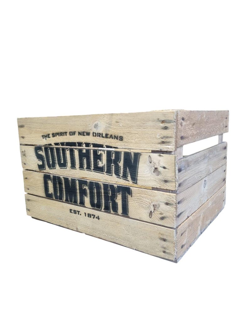 Pode incluir: Uma caixa de madeira r&uacute;stica com as palavras "SOUTHERN COMFORT" em letras pretas grandes. O texto "THE SPIRIT OF NEW ORLEANS" e "EST. 1874" tamb&eacute;m s&atilde;o vis&iacute;veis. A caixa &eacute; feita de madeira clara e tem formato retangular.