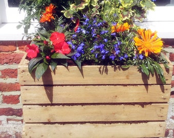 Op de afbeelding: Een houten plantenbak met een verscheidenheid aan bloemen, waaronder blauwe, rode en oranje bloesems. De plantenbak is gemaakt van natuurlijk hout en heeft een rustieke uitstraling.