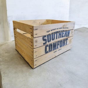 Pode incluir: Caixa de madeira com o texto "SOUTHERN COMFORT" em letras pretas. A caixa &eacute; feita de madeira clara e tem formato retangular com ripas. O texto "THE SPIRIT OF NEW ORLEANS" e "EST. 1874" tamb&eacute;m s&atilde;o vis&iacute;veis.