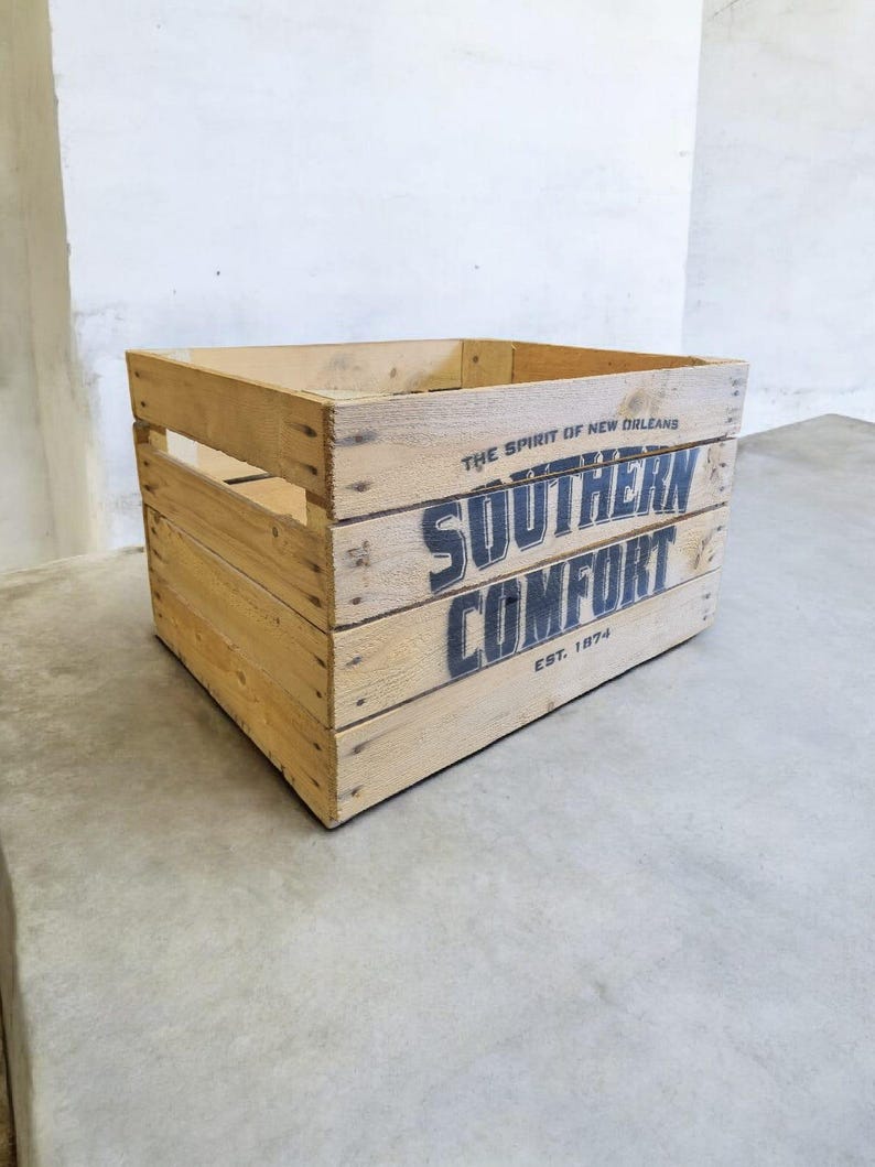 Pode incluir: Caixa de madeira com o texto "SOUTHERN COMFORT" em letras pretas. A caixa &eacute; feita de madeira clara e tem formato retangular com ripas. O texto "THE SPIRIT OF NEW ORLEANS" e "EST. 1874" tamb&eacute;m s&atilde;o vis&iacute;veis.