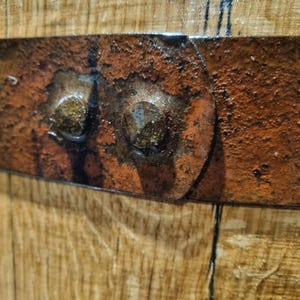 May include: Close-up of a section of a wooden barrel. The image shows the aged wood grain and a rusty metal band secured with two large, weathered bolts. The metal band is a deep reddish-brown color, contrasting with the lighter wood.
