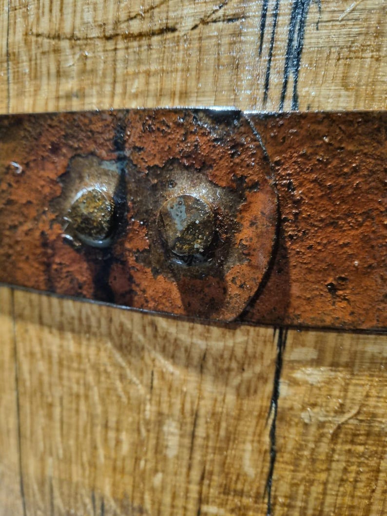 May include: Close-up of a section of a wooden barrel. The image shows the aged wood grain and a rusty metal band secured with two large, weathered bolts. The metal band is a deep reddish-brown color, contrasting with the lighter wood.