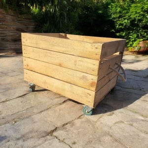 Può includere: Una cassa di legno rettangolare con finitura naturale, montata su quattro piccole ruote blu. La cassa ha maniglie in corda sui lati ed è costruita con assi di legno orizzontali. La cassa è all'aperto su una superficie di pietra.