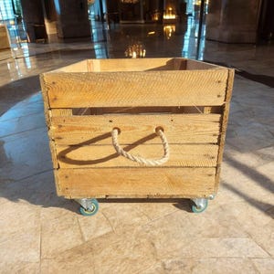 Peut inclure: Caisse en bois naturel avec une poignée en corde et quatre petites roulettes bleues. La caisse est fabriquée à partir de lattes de bois horizontales. Idéale pour le stockage ou l'exposition.