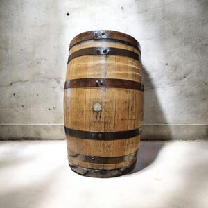 May include: A wooden barrel with metal bands. The barrel is made of light brown wood and has several dark metal bands around its circumference. A small, round wooden plug is visible on the front. The barrel is standing in front of a textured gray wall.