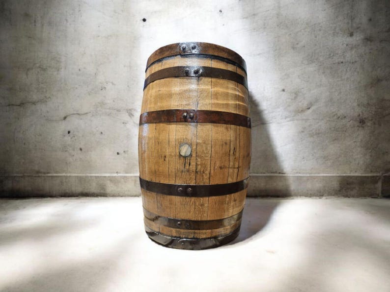 May include: A wooden barrel with metal bands. The barrel is made of light brown wood and has several dark metal bands around its circumference. A small, round wooden plug is visible on the front. The barrel is standing in front of a textured gray wall.