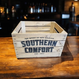 Pode incluir: Caixa de madeira com as palavras "SOUTHERN COMFORT" em letras pretas. A caixa tamb&eacute;m tem o texto "THE SPIRIT OF NEW ORLEANS" e "EST. 1874". Est&aacute; sobre uma superf&iacute;cie de madeira.