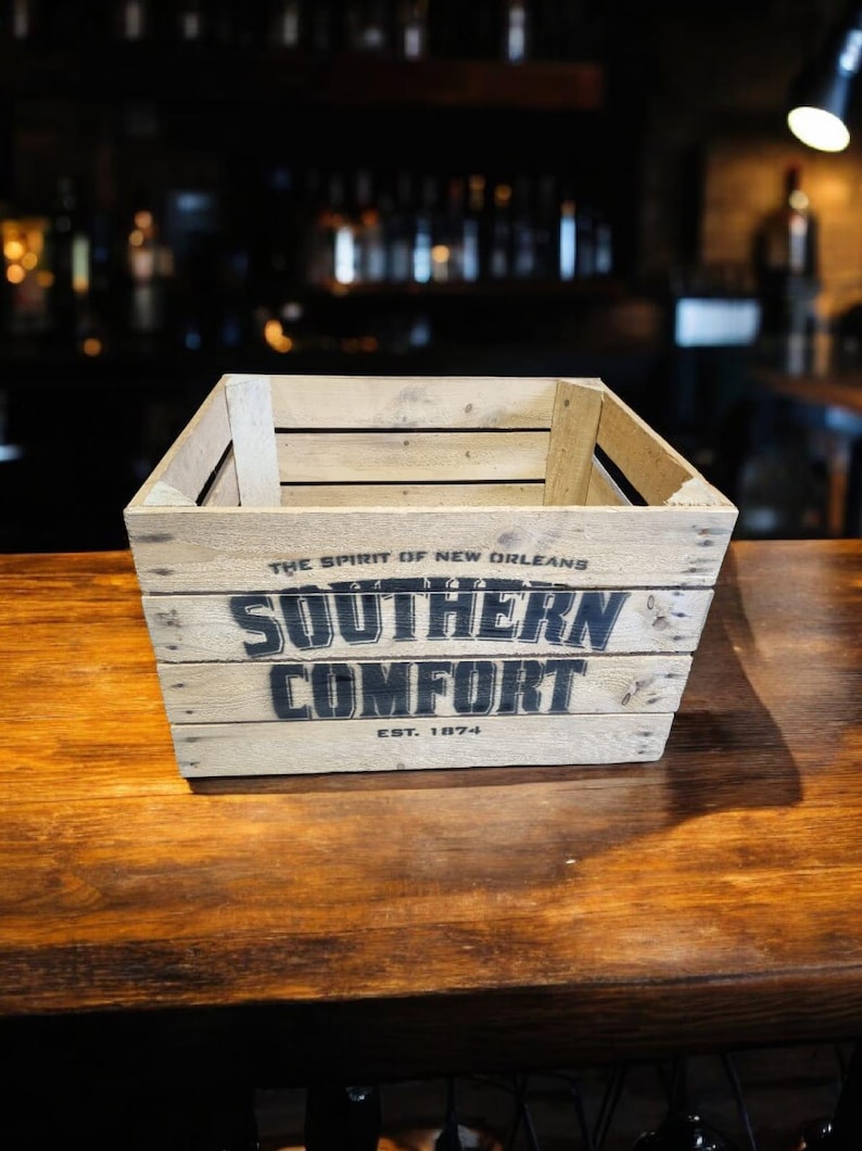 Pode incluir: Caixa de madeira com as palavras "SOUTHERN COMFORT" em letras pretas. A caixa tamb&eacute;m tem o texto "THE SPIRIT OF NEW ORLEANS" e "EST. 1874". Est&aacute; sobre uma superf&iacute;cie de madeira.