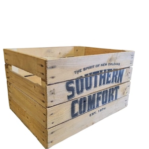 Pode incluir: Uma caixa de madeira com as palavras "SOUTHERN COMFORT" em letras grandes, azul escuro. O texto "THE SPIRIT OF NEW ORLEANS" e "EST. 1874" tamb&eacute;m s&atilde;o vis&iacute;veis. A caixa &eacute; feita de madeira clara.