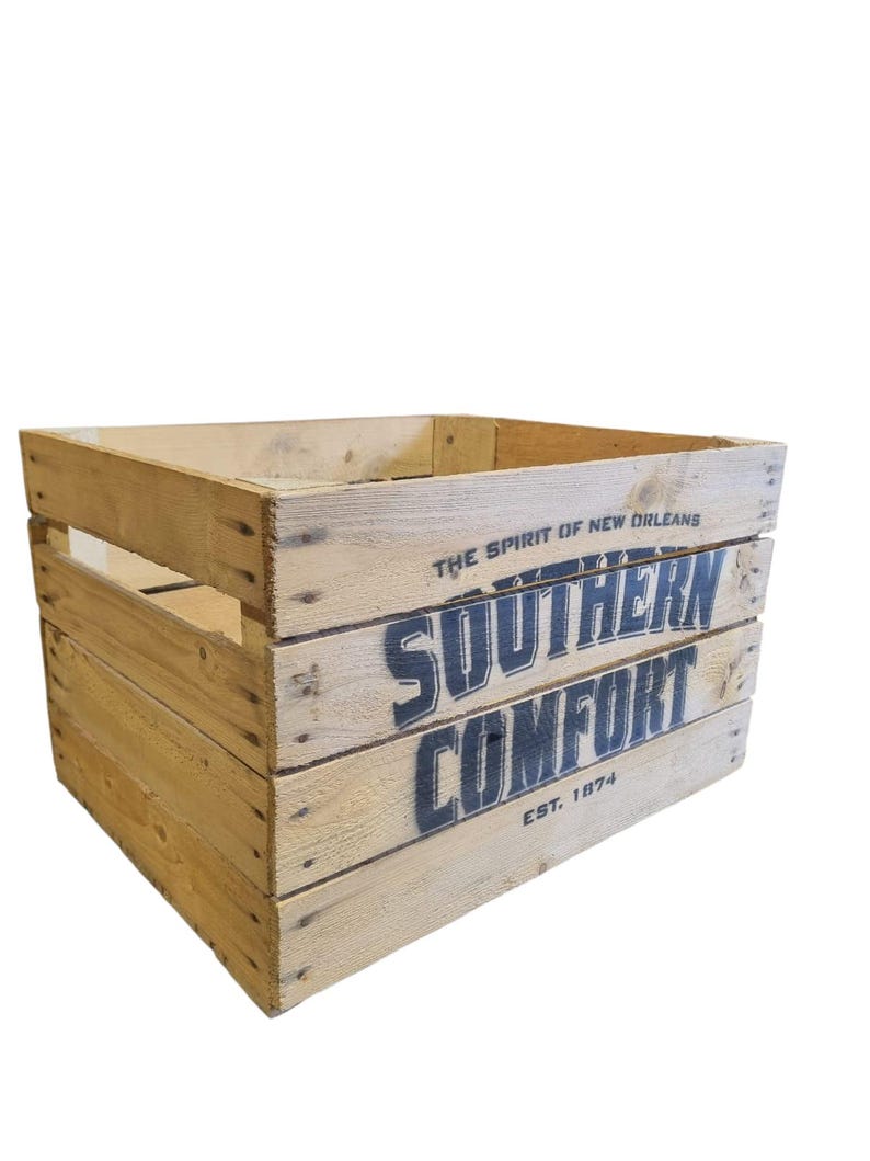 Pode incluir: Uma caixa de madeira com as palavras "SOUTHERN COMFORT" em letras grandes, azul escuro. O texto "THE SPIRIT OF NEW ORLEANS" e "EST. 1874" tamb&eacute;m s&atilde;o vis&iacute;veis. A caixa &eacute; feita de madeira clara.