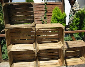 May include: A stack of six rustic wooden crates, arranged in a staggered formation. The crates are constructed from weathered, light brown wood with visible grain and slats. The scene is outdoors, with greenery in the background.