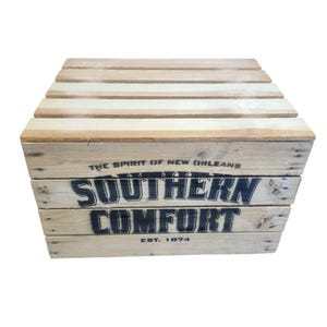 Pode incluir: Uma caixa de madeira com o texto "SOUTHERN COMFORT" em letras pretas. A caixa &eacute; feita de madeira clara e tem um tampo de ripas. O texto "EST. 1874" tamb&eacute;m &eacute; vis&iacute;vel. As palavras "THE SPIRIT OF NEW ORLEANS" s&atilde;o impressas acima do texto principal.