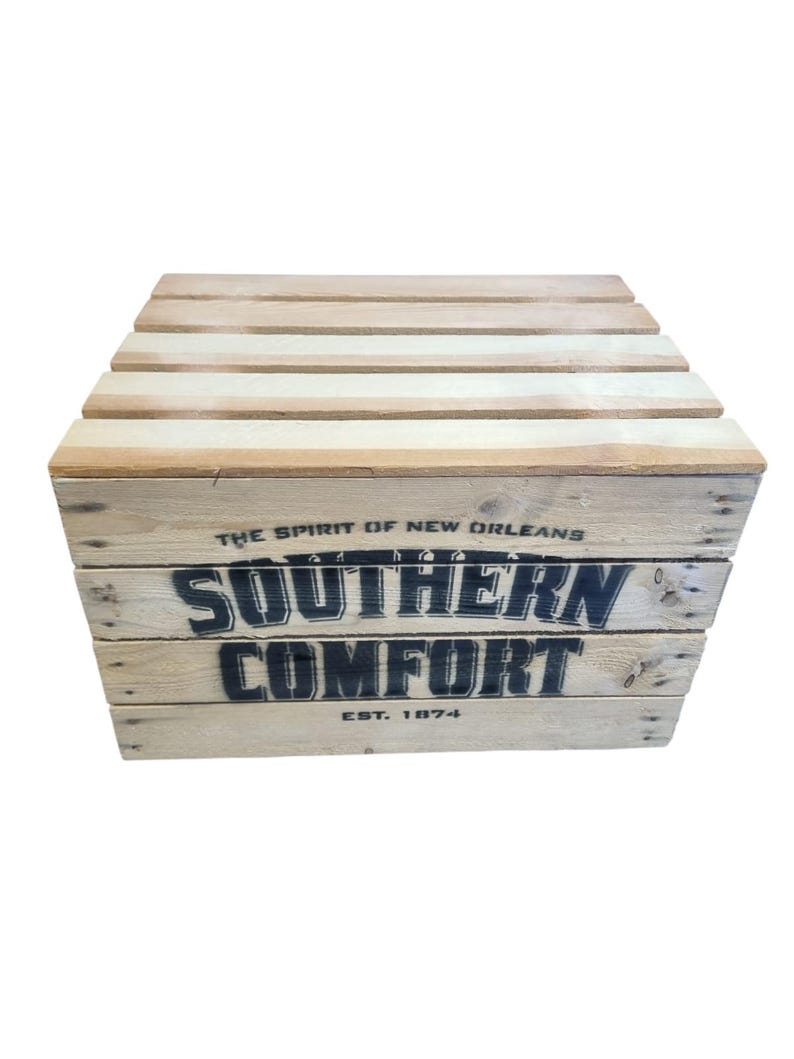 Pode incluir: Uma caixa de madeira com o texto "SOUTHERN COMFORT" em letras pretas. A caixa &eacute; feita de madeira clara e tem um tampo de ripas. O texto "EST. 1874" tamb&eacute;m &eacute; vis&iacute;vel. As palavras "THE SPIRIT OF NEW ORLEANS" s&atilde;o impressas acima do texto principal.