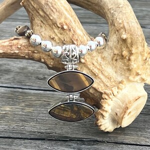 Pode incluir: Um bracelete de contas de prata com um pingente de dupla face com pedra de olho de tigre. O pingente tem formato oval e um design de filigrana de prata.