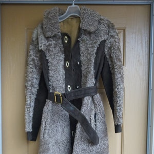 May include: A vintage, knee-length coat with a sheepskin-like texture. The coat features a dark brown suede trim, a matching belt with a gold-colored buckle, and a button-up front. The coat is hanging on a white hanger against a light brown door.