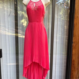 Puede incluir: Un vestido rosa brillante con cuello halter, falda plisada y dobladillo asim&eacute;trico. El corpi&ntilde;o presenta un detalle de ojo de cerradura y detalles de encaje. El vestido se exhibe en un maniqu&iacute;.