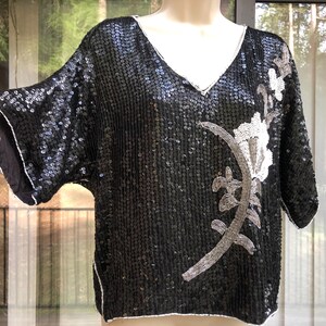 May include: Black sequined top with a V-neck and short sleeves. The top features a floral design on the front, with white and silver beaded accents. The edges are trimmed with white piping.