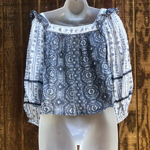Puede incluir: Una blusa estampada en azul y blanco con mangas largas y cuello cuadrado. La blusa presenta un dise&ntilde;o geom&eacute;trico en la parte delantera y estampados florales en las mangas. Se exhibe en un maniqu&iacute; blanco.