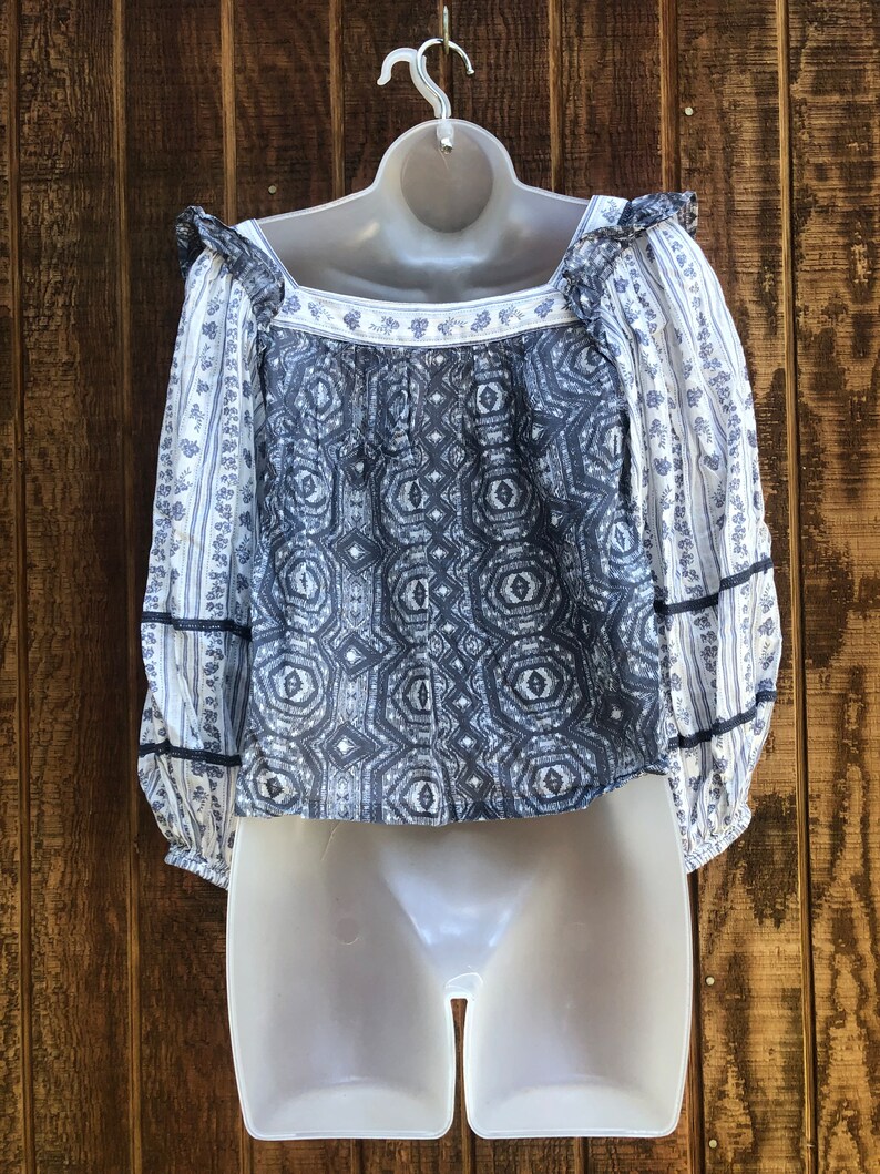 Puede incluir: Una blusa estampada en azul y blanco con mangas largas y cuello cuadrado. La blusa presenta un dise&ntilde;o geom&eacute;trico en la parte delantera y estampados florales en las mangas. Se exhibe en un maniqu&iacute; blanco.