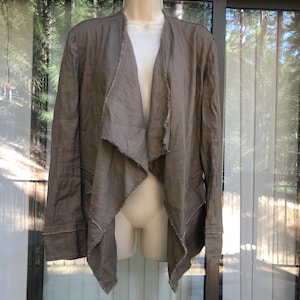 K&ouml;nnte beinhalten: Taupefarbene Jacke mit offenem Schnitt, drapiertem Kragen und langen &Auml;rmeln. Die Jacke hat rohe Kanten am Kragen, an den Manschetten und am Saum. Die Jacke wird auf einer Schaufensterpuppe vor einem Fenster pr&auml;sentiert.