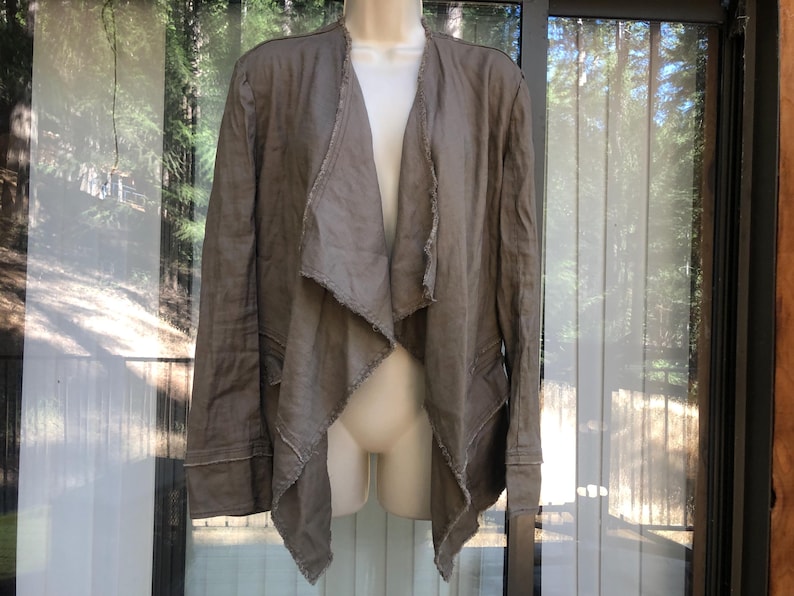 K&ouml;nnte beinhalten: Taupefarbene Jacke mit offenem Schnitt, drapiertem Kragen und langen &Auml;rmeln. Die Jacke hat rohe Kanten am Kragen, an den Manschetten und am Saum. Die Jacke wird auf einer Schaufensterpuppe vor einem Fenster pr&auml;sentiert.
